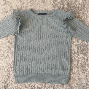 Ljusgrå stickad tröja med volang - Supersöt grön stickad tröja från XOXO friends med volangdetaljer på axlarna. Tröjan har kabelstickat mönster, rund halsringning och långa ärmar. Perfekt för dig som gillar en söt och trendig look.