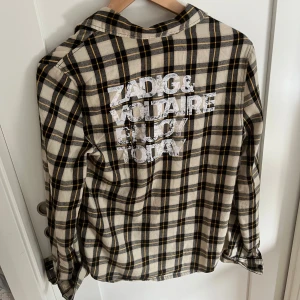 Rutig skjorta från Zadig & Voltaire - Snygg rutig skjorta från Zadig & Voltaire i beige, och svart. Skjortan har tryckt text på ryggen och klassisk krage. Tillverkad i mjuk bomull och har långa ärmar med knappar vid ärmslut. Perfekt för en cool och avslappnad stil.