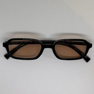 Svarta rektangulära solglasögon - Snygga solglasögon med svart, glansig båge och rektangulär form. Glasen är bruna och ger en cool retrovibe. Perfekta för dig som vill sticka ut med en stilren accessoar. Passar till de flesta outfits och ger en trendig look.