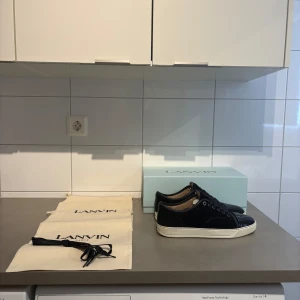 Svarta sneakers från Lanvin i mocka - Snygga navyblåa sneakers från Lanvin med ovandel i mocka och lackad tå. Skorna har vita platta sulor och svarta snören. Klassisk och stilren design med exklusiv känsla, perfekt för dig som gillar premium streetstyle. Har massa mer bilder. Använt sparsamt, säljer pga försmå. Köpta från care of Carl för 4 500kr. Storlek UK 9