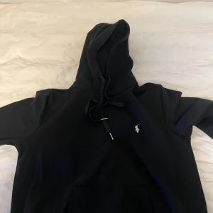 Svart hoodie från Polo Ralph Lauren - Svart hoodie från Polo Ralph Lauren med klassisk broderad logga på bröstet. Tröjan har huva med snörning, känguruficka och lång ärm. Mjuk bomullskvalitet och relaxed fit – perfekt för chill dagar eller när du vill ha en clean look. Använt en skoldag bara, den var för liten i armarna.