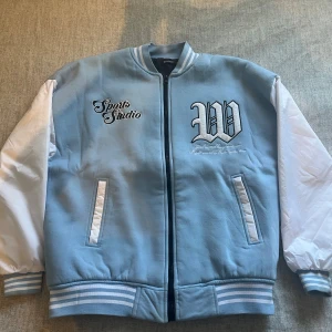 Blåvit bomberjacka Sports Studio - Snygg bomberjacka i ljusblått med vita ärmar och broderade detaljer framtill. Säljer jackan för att den inte passar mig längre. Priset kan diskuteras vid snabb affär!