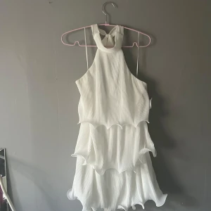 Vit volangklänning från Gina Tricot - Supersöt vit kortklänning från Gina Tricot med flera lager volanger och plisserat tyg. Klänningen har halterneck med knyt i nacken och är ärmlös. Perfekt för sommarens alla tillfällen när du vill sticka ut lite extra.