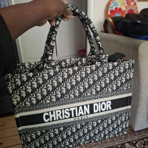 Christian Dior Book Tote axelväska - Säljer en ikonisk Christian Dior Book Tote axelväska i svart och beige med klassiskt Dior-mönster. Väskan är rymlig med två stadiga handtag och har ett stort öppet fack samt innerficka med dragkedja. Perfekt för dig som vill ha en statementväska med lyxig känsla.