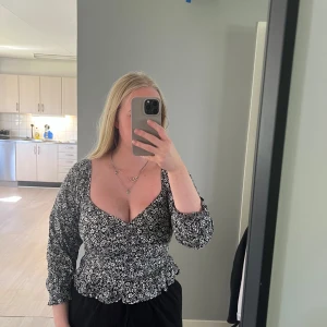 Svart blommig blus med volang - Säljer en svart blus med vita små blommor, v-ringad och med långärmade puffärmar. Blusen har en smockad midja och volangkant nedtill.