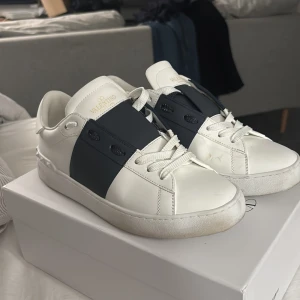 Valentino Garavani Open sneakers vit/svart - Snygga Valentino Garavani Open sneakers i vitt skinn med bred svart rem över mitten. Klassisk rund tå, vita snören och diskret guldlogga på plösen. Kommer med originalpåse och box – perfekt för dig som gillar exklusiva streetwear-vibbar.