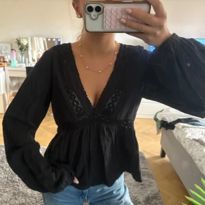 Svart blus med spets och v-ringning - Säljer en svart blus med djup v-ringning och snygga spetsdetaljer längs halsen och midjan. Blusen har långa, lite puffiga ärmar med resår fram vid handen. Den är använd 1 gång så toppen skick, som ny. Storlek xs men passar S också. 