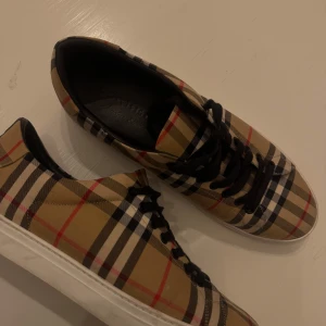 Burberry sneakers  - Snygga sneakers från Burberry i klassiskt beige, svart, vit och röd rutig design. Skorna har svarta snören, vit platt sula och insida i svart skinn. Perfekta för dig som vill sticka ut med en mer grich stil. Dom är väldigt svåra att få tag på marknaden.