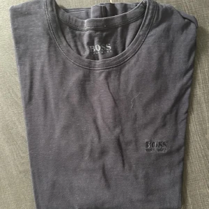 Svart t-shirt från Hugo Boss - Svart t-shirt från Hugo Boss i mjuk bomull. Klassisk rund hals och korta ärmar. Diskret BOSS-logga broderad på bröstet. Passformen är normal och t-shirten är enkel att matcha med jeans eller shorts. Perfekt för dig som gillar stilrena och snygga basplagg.