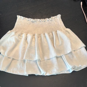 Ljusvit volangkjol med smock - Supersöt kort kjol i ljusvit nyans med bred smock i midjan och dubbla volanger. Kjolen har en flowy känsla och är perfekt för varma dagar. Materialet känns lätt och luftigt, vilket gör den extra bekväm att bära.