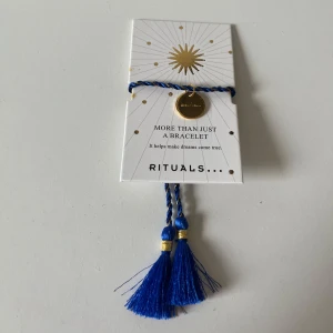 Blått armband från Rituals med tofsar - Snyggt armband från Rituals i blått och guld med tvinnad design och två stora tofsar. Armbandet har en rund guldfärgad berlock med Rituals-loggan och detaljer i guld. 