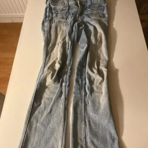 Bootcut jeans i ljusblå denim från H&M - Ljusblå bootcut jeans från H&M med låg midja och justerbar midja. Klassisk femficksmodell med snygga ficklock och silverfärgade knappar framtill. Hål vid grenen.