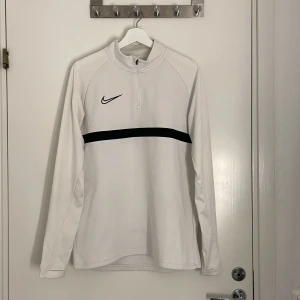 Vit Nike Dri-Fit half zip tröja - Nike Dri-Fit tröja i vitt med svart rand över bröstet och svart Nike-logga. Långärmad modell med half zip och hög krage. Perfekt för träning eller chill dagar.