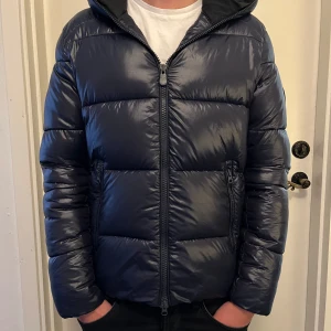  Save The Duck jacka  - Säljer en mörkblå pufferjacka från Save The Duck med huva och dragkedja framtill. Jackan är vadderad med syntetiskt dun (Plumtech) och har två fickor med dragkedja. Perfekt för kalla dagar och riktigt skön att bära. Skicket är 9/10,storlek S, kan gå ner i pris vid snabb affär 