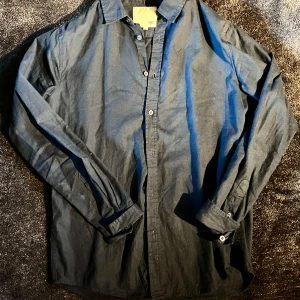 Mörkblå linneskjorta med knappar - En stilren mörkblå skjorta från 157 i mjuk linne. Skjortan har klassisk krage, långa ärmar och knäppning framtill. Passar perfekt till jeans eller chinos för en clean look. Enkel och tidlös design som funkar året runt.