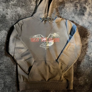 Svart hoodie med örntryck och snörning - Svart hoodie från Modern Essentials med fett örntryck och röd text 'Only The Brave' på bröstet. Tröjan har huva med snörning, stor magficka och ribbade muddar. Perfekt för dig som gillar streetwear och vill sticka ut lite extra.