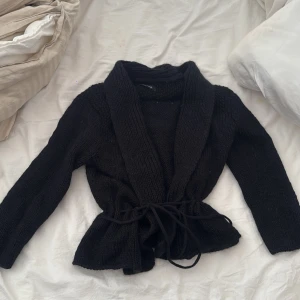 Svart stickad kofta från Samsøe Samsøe - Svart, chunky stickad kofta från Samsøe Samsøe med bred krage och knytband i midjan. Koftan har långärmade ärmar och en loose passform som ger en chill vibe. Perfekt att slänga på sig när det är kallt ute.