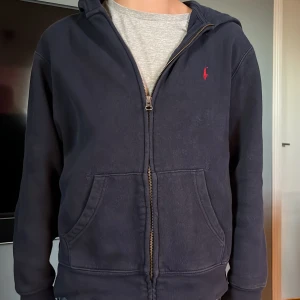 Polo Ralph Lauren zip hoddie  - Säljer en mörkblå hoodie från Polo Ralph Lauren med dragkedja och huva. Tröjan har en röd broderad logga på bröstet. Perfekt för chill dagar och enkel att matcha med jeans eller joggers.