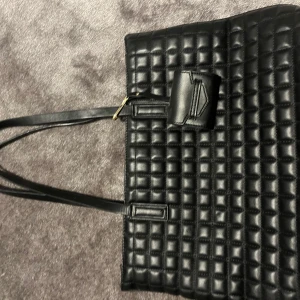 Svart quiltad axelväska i skinnimitation - Snygg svart axelväska med quiltad design och långa axelband. Väskan har ett stort huvudfack, guldfärgade detaljer och dragkedja upptill. Tillverkad i skinnimitation och perfekt för dig som vill ha en stilren och rymlig väska med modern känsla.