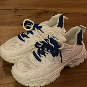 Chunky vita sneakers med blå detaljer - Snygga chunky sneakers i vitt med blå snörning och blå detaljer vid häl och plös. Ovandelen har meshpaneler och syntetiska partier för en sportig look. Grov sula ger extra höjd och streetkänsla. Perfekta för dig som gillar trendiga och bekväma skor.
