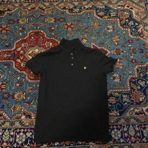 Svart pikétröja från Lyle & Scott - Klassisk svart pikétröja från Lyle & Scott med korta ärmar och gul logga på bröstet. Tröjan har krage och knäppning med tre knappar framtill. Perfekt för en stilren och enkel look.