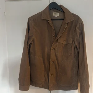 Brun manchester overshirt från East West - Säljer en brun overshirt från East West i mjuk manchester. Jackan har klassisk krage, tre fickor framtill och knäppning med knappar. Perfekt att slänga över en hoodie eller t-shirt för en chill look.