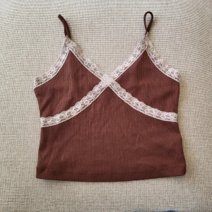 Brun ribbad linnetopp med spets - Supersöt brun ribbad linnetopp med tunna axelband och vit spetsdetalj längs urringningen och över bysten. Mjuk och stretchig känsla. Lite nopprig men inget som  jag anser förstör helhetsintrycket. 