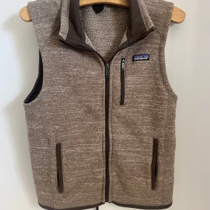 Brun fleeceväst från Patagonia - Mysig brun fleeceväst från Patagonia med hög krage och hel dragkedja framtill. Västen har tre fickor med dragkedja och ett diskret melerat mönster. Perfekt för lager-på-lager och chill höst- eller vinterdagar.