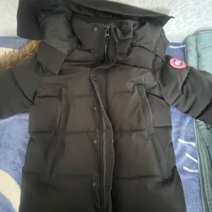 Svart dunjacka från Canada Goose - Säljer en svart dunjacka från Canada Goose med klassisk patch på ärmen. Jackan har stor huva med avtagbar fuskpäls, dragkedja och knappar framtill samt två stora fickor. Perfekt för kalla vinterdagar och riktigt snygg streetstil. Den passar också om du tar på dig M 