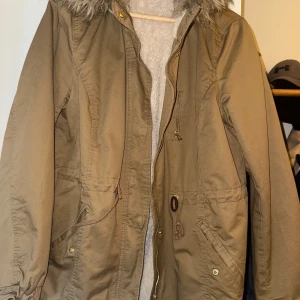 Beige/khaki parkajacka med fuskpäls från H&M - Parkajacka i beige från H&M med mysig fuskpäls i luvan och dragkedja framtill. Jackan har snörning i midjan, fickor med knappar och är fodrad med mjukt material. Perfekt för kyliga dagar och har en klassisk, avslappnad look.