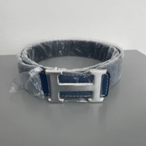  bälte i blått och svart skinn - Snyggt  bälte i blått och svart skinn med ikoniskt H-spänne i silverfärgad metall. Bältet har stilrena kontrastsömmar och klassisk design som lyfter vilken outfit som helst. Perfekt accessoar för dig som gillar exklusiva detaljer och tidlös stil.
