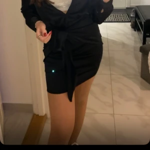 Svart klänning från Bik Bok - Snygg svart kortklänning från Bik Bok i satinliknande material. Klänningen har långa ärmar, knappar framtill och en stor knytning i midjan.