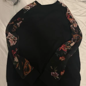 Svart sweatshirt med blommiga ärmar - Svart sweatshirt med långa ärmar som har ett färgstarkt blommigt mönster längs hela ärmarna. Tröjan har en klassisk rund halsringning och är perfekt för dig som vill ha en stilren look med en twist. Skön och mjuk, passar till många outfits. Passar även bra nu under hösten!!