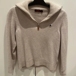 Beige stickad tröja från Morris - Snygg beige stickad tröja från Morris med ribbad krage och half zip-dragkedja. Tröjan har långärmad design och en broderad svartvit logga på bröstet. Perfekt för lager-på-lager och chill höst- eller vinterstil.