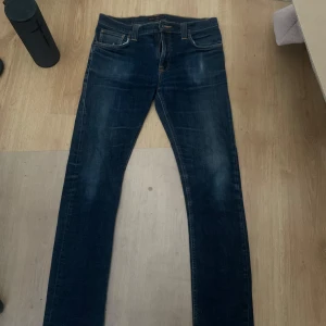 Mörkblå jeans från Nudie Jeans W33 L34 - Snygga mörkblå jeans från Nudie Jeans med klassiska orange sömmar och broderade bakfickor. Modellen har en rak passform och är tillverkad i slitstarkt denim. Perfekta för dig som gillar stilrena och tidlösa jeans.