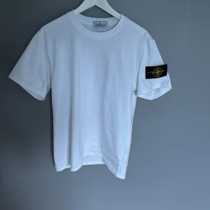 Vit Stone Island t-shirt - Vit t-shirt från Stone Island med klassisk rund hals och korta ärmar. På vänster ärm sitter den ikoniska svarta patchen med gult och grönt broderi. Tillverkad i mjuk bomull för en skön känsla och clean look. Den har bara blivit hängande i garderoben, använd max 3 gånger, size M, nyskick