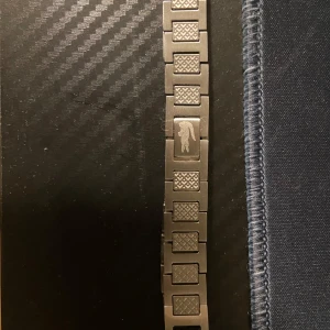 Silverfärgat armband med mönster - Stilrent armband i silverfärgad metall med rektangulära länkar och ett rutigt mönster på varje länk. På en av länkarna finns en ingraverad panter, vilket ger en cool och unik touch. Perfekt för dig som gillar detaljer och vill sticka ut.