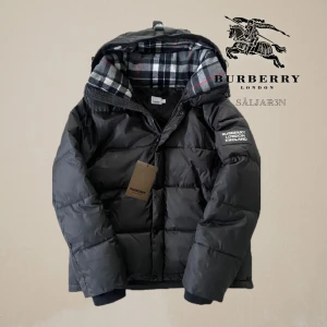 Burberry Jacka - Säljer en exklusiv Burberry dunjacka i svart, storlek M/L, i nyskick med taggar. Modellen är mångsidig med 2-i-1 funktion – ärmarna kan tas av så att jackan blir en stilren väst, perfekt året runt. Dunfodret ger värme och komfort utan att kännas tungt. Avtagbar huva med klassiskt rutmönster, dragkedjeförsedda fickor samt diskret patch på ärmen. En tidlös premiumjacka som passar både till vardag och mer stilfulla tillfällen. 📦Snabb leverans! - Skickas samma dag!📦