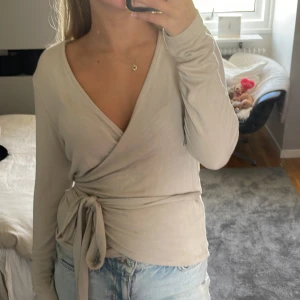 Beige omlottopp med knytband - Supersnygg beige omlottopp från Lager 157 med långa ärmar och djup v-ringning. Toppen har ett brett knytband i midjan som ger en riktigt schysst siluett. Perfekt att styla med jeans eller kjol för en chill men trendig look. Endast använd nån gång.
