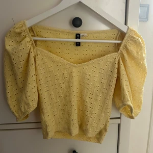Gul blus med puffärm från Divided - Söt gul blus från Divided med broderade hål och puffiga korta ärmar. Toppen har en v-ringad hals och elastiska detaljer vid axlarna för en trendig look. Perfekt för varma dagar när du vill sticka ut lite extra.