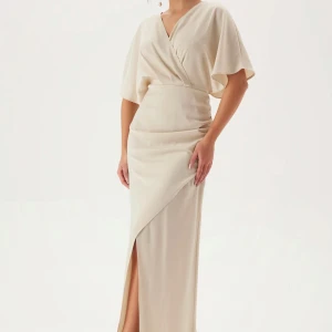 Champagnefärgad omlottklänning med slits - Säljer en elegant långklänning i champagnefärgad ton med omlottdesign och v-ringad hals. Klänningen har korta, vida ärmar och en snygg slits framtill som ger extra edge. Använd vid ett tillfälle inga defekter!!