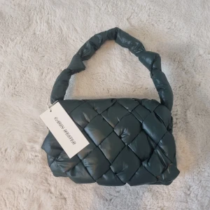 Mörkgrön vadderad axelväska Carin Wester - Säljer en mörkgrön vadderad axelväska från Carin Wester. Väskan har en chunky, flätad design och ett mjukt, puffigt handtag. Tillverkad i syntetmaterial och perfekt för dig som vill ha en cool och trendig accessoar med unik känsla.