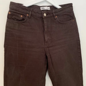 Bruna raka jeans från Zara - Bruna jeans från Zara med klassisk femficksdesign och raka ben. Byxorna har medium/hög midja och slitsar nertill vid bensluten för en snygg detalj. Tillverkade i bomull med en skön känsla och stilren look.