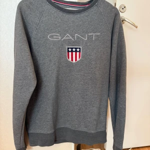 Grå sweatshirt från GANT - Snygg grå sweatshirt från GANT med broderad logga och emblem på bröstet. Tröjan har rund hals, långa ärmar och ribbade muddar. Perfekt för dig som gillar klassisk och sportig stil med en amerikansk touch.