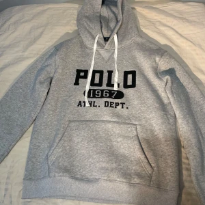Grå hoodie från Polo Ralph Lauren - Snygg grå hoodie från Polo Ralph Lauren med svart tryck framtill, stor huva och vita snören. Klassisk känguruficka och ribbade muddar. Perfekt för chill dagar och streetstyle. Mjuk bomullsblandning som känns skön mot huden.