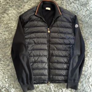 Svart dunjacka från Moncler - Snygg svart dunjacka från Moncler med quiltad framsida och släta, stickade ärmar. Jackan har dragkedja framtill, ribbad krage med röd och vit kant, samt Moncler-logga på ärmen. Perfekt för dig som vill ha en stilren och sportig look.