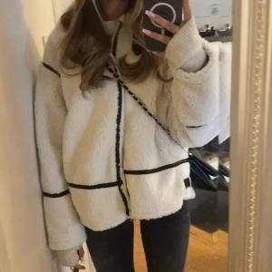 Vit teddyjacka med svarta detaljer - Supermysig vit teddyjacka med svarta linjer som ger en cool kontrast. Jackan har oversized passform, dragkedja framtill och långa ärmar. Perfekt för att hålla sig varm och ändå se trendig ut.