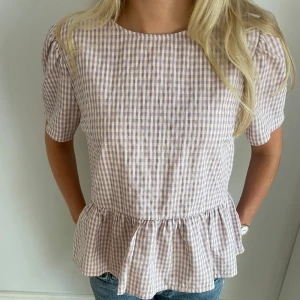 Ljuslila rutig blus med rosetter - Supersöt blus i ljuslila och vitt rutigt mönster. Toppen har korta puffärmar, volang nertill och öppen rygg med två stora knytrosetter. Perfekt för dig som gillar romantiska detaljer och en feminin stil.