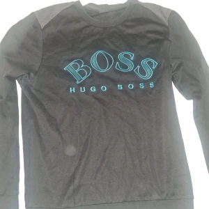 Svart tröja från Hugo Boss med logga - Svart långärmad tröja från Hugo Boss med stor BOSS-logga broderad på bröstet. Tröjan har rund halsringning, ribbade muddar och detaljer på axlarna. 