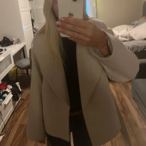 Beige omlottkappa med bälte - Säljer en snygg beige kappa i omlottmodell med bälte i midjan. Jackan har en loose passform och stora slag, perfekt för lager-på-lager. Materialet känns mjukt och ger en clean look. Passar dig som gillar stilrena och enkla plagg.